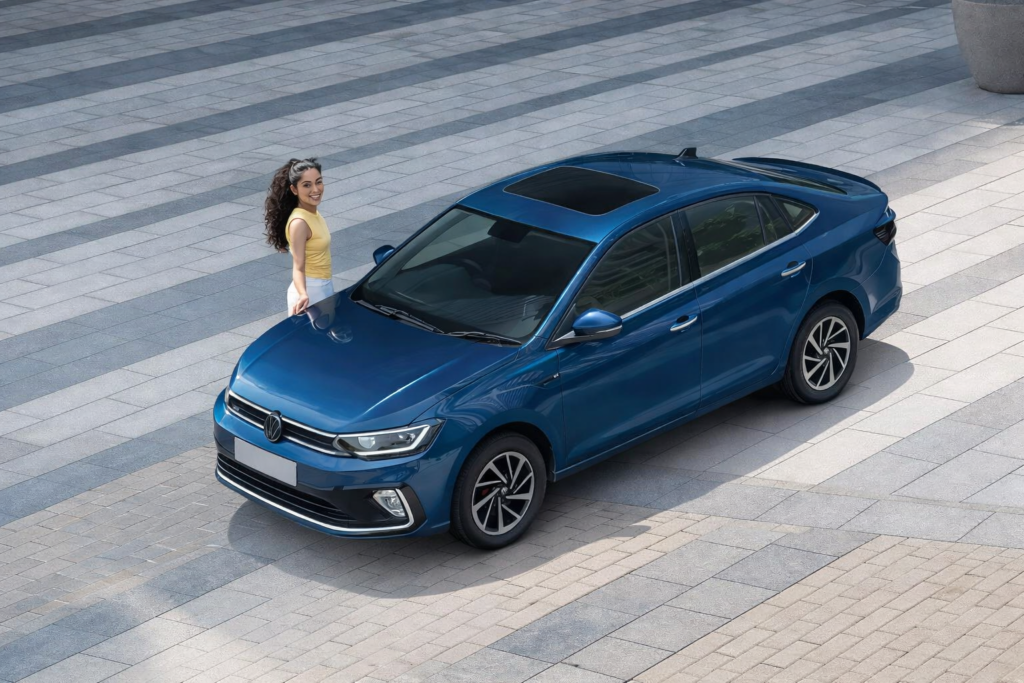 Blue Volkswagen Virtus sedan parked in an open modern plaza with a woman standing beside the car, showcasing its sleek design and alloy wheels.