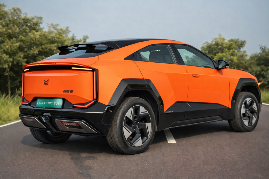 Orange Mahindra BE 6 electric SUV rear three-quarter view with sharp LED tail lights, coupe-style design, black cladding, and sporty alloy wheels parked on an open road.