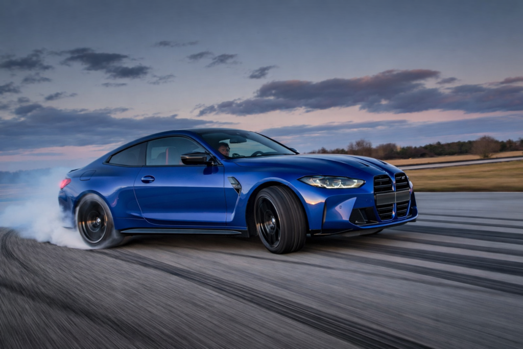 A blue BMW M4 sports coupe drifting on a racetrack at dusk, with rear tires producing smoke, aggressive front grille visible, and motion blur on the road surface.