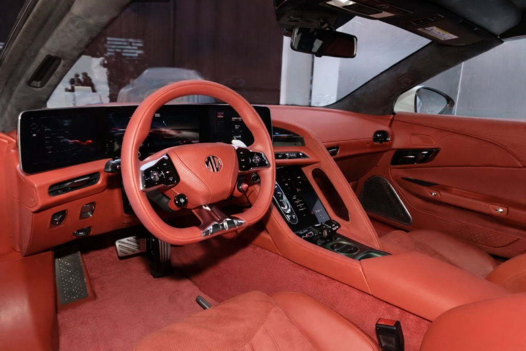 Orange leather interior of a modern sports car featuring a digital instrument cluster, large touchscreen display, and a sporty multi-function steering wheel.