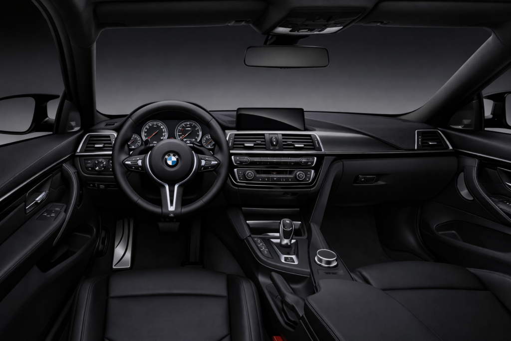 Interior view of a BMW M4 sports coupe showing a black leather dashboard, steering wheel with BMW logo, digital instrument cluster, center console, and a clean modern cockpit design.